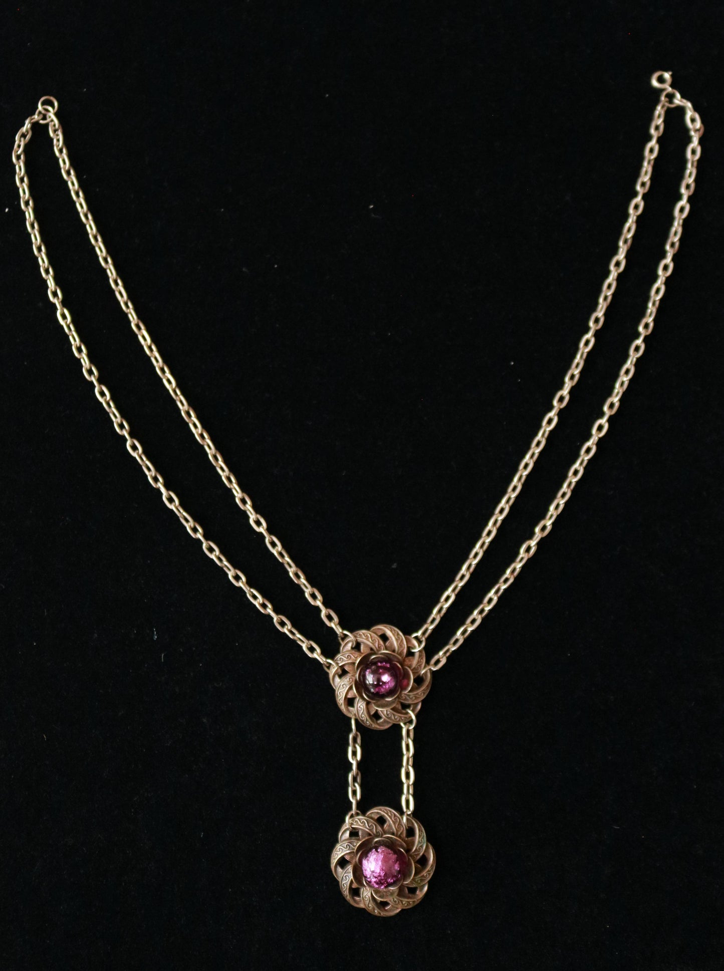 Vintage floral drop necklace with two ornate metal flower motifs set with purple Murano glass cabochons on a black velvet background.